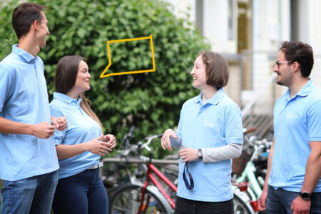 Vier Studierende mit einheitlichen hellblauen Shirts stehen vor einem grünen Hintergrund und unterhalten sich