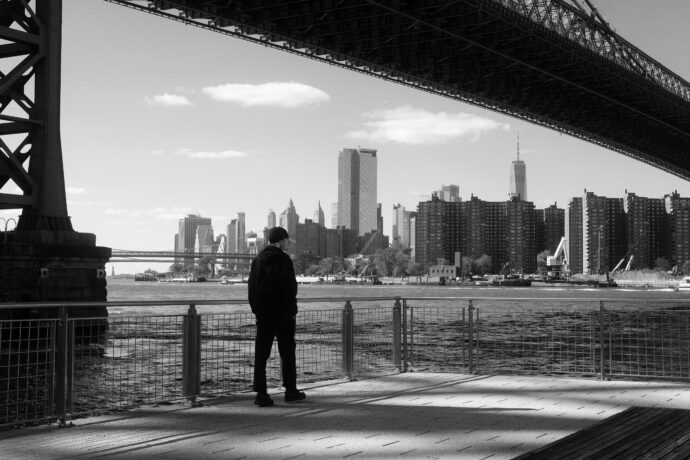 Das Schwarz-Weiß-Bild zeigt einen Mann, der in New york unter einer Brücke steht und in die Ferne guckt.