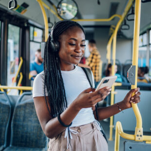 Eine junge Frau mit Kopfhörern steht in einem Bus, hält sich an einer Stange fest und schaut lächelnd auf ihr Smartphone. Im Hintergrund sitzen und stehen weitere Fahrgäste.