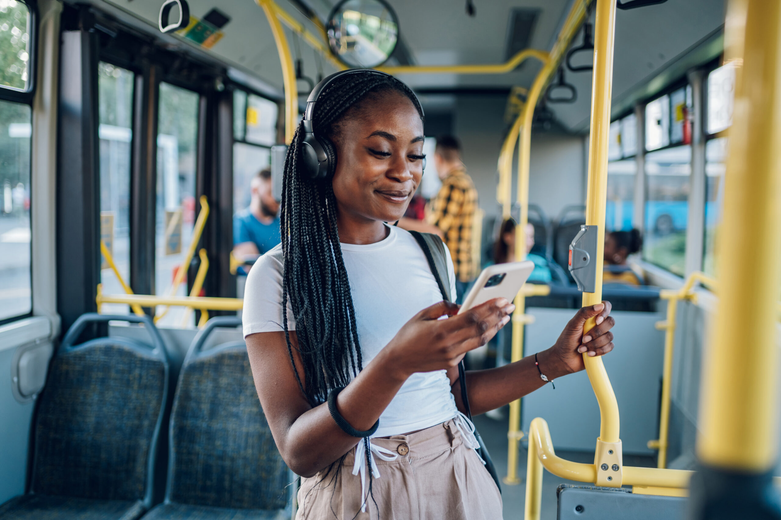 Eine junge Frau mit Kopfhörern steht in einem Bus, hält sich an einer Stange fest und schaut lächelnd auf ihr Smartphone. Im Hintergrund sitzen und stehen weitere Fahrgäste.